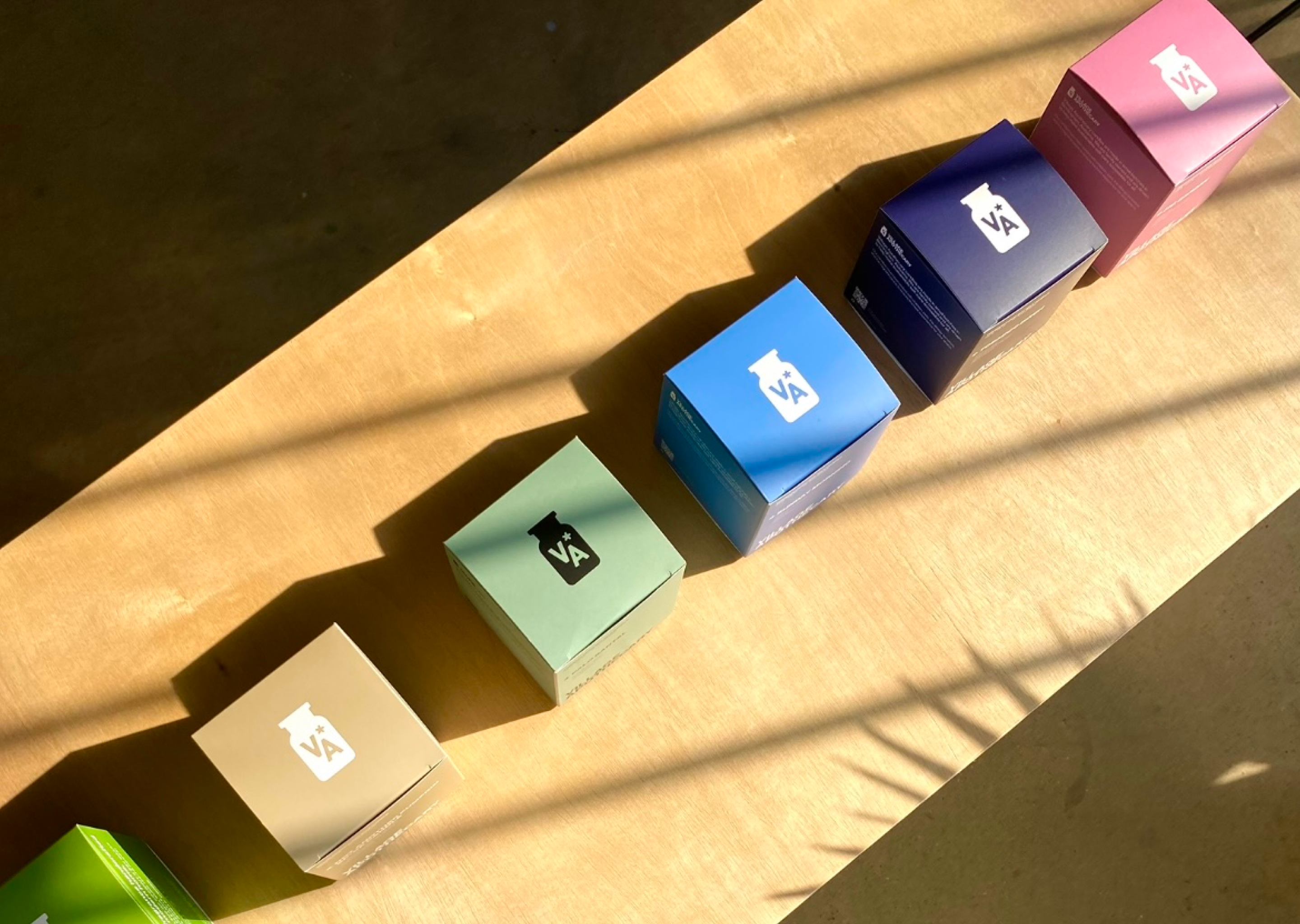 Six small branded product boxes arranged diagonally on a sunlit wooden surface. The boxes come in beige, green, blue, navy, pink, and a partially visible green box, each with a white logo.